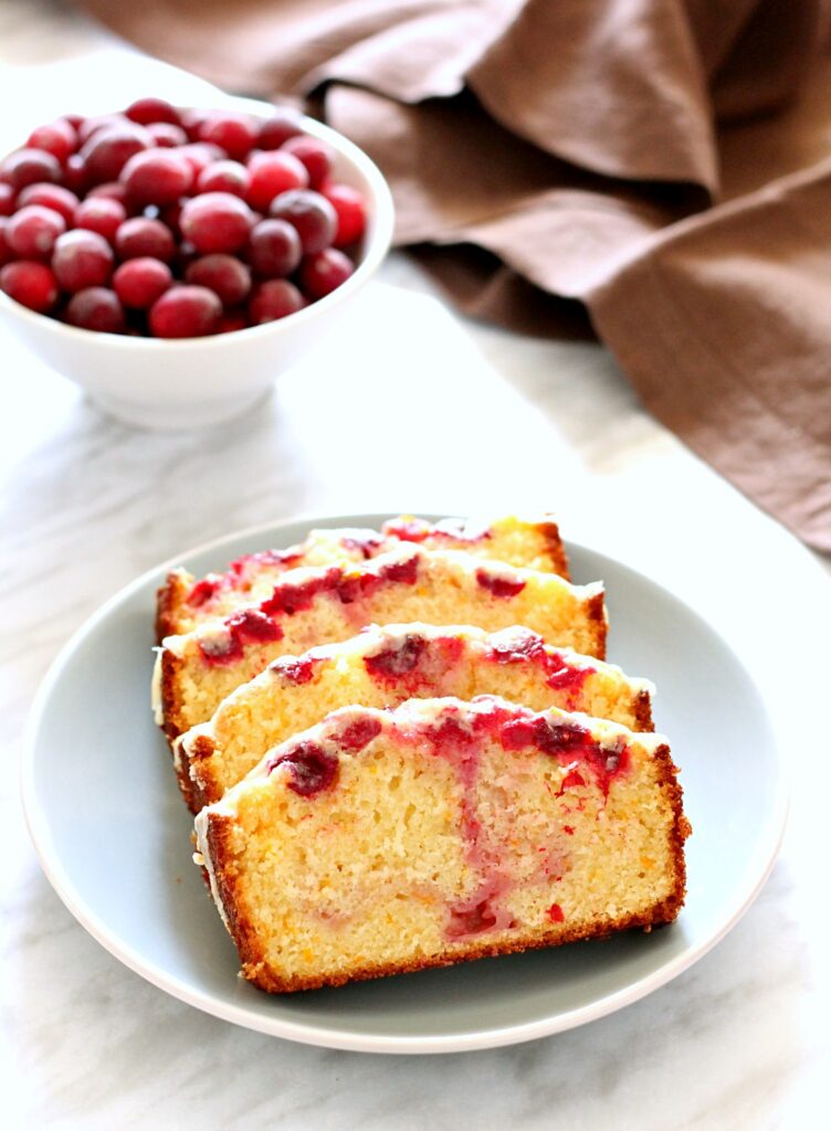 Four slices of Cranberry Orange Loaf on a blue plate with a bowl of cranberries in the background.
