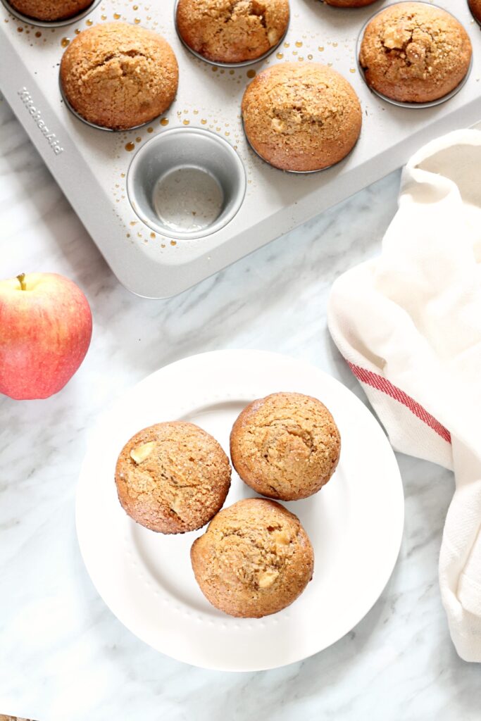 Three Whole Wheat Apple Muffins on a white plate beside a muffin tin.